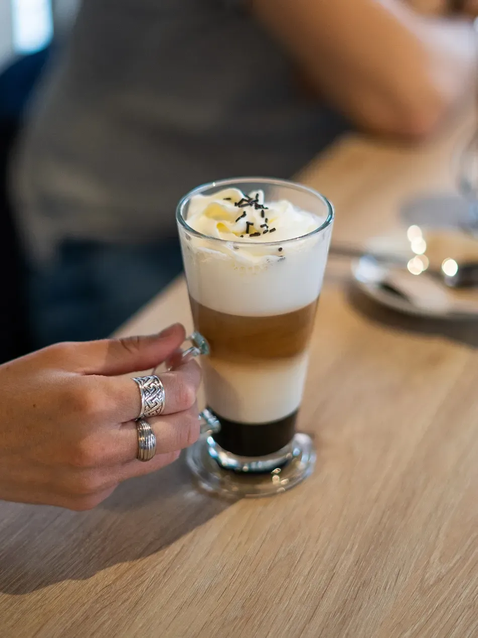 Una mano sostiene una bebida de café con varias capas de espuma sobre una mesa de madera en una cafetería.