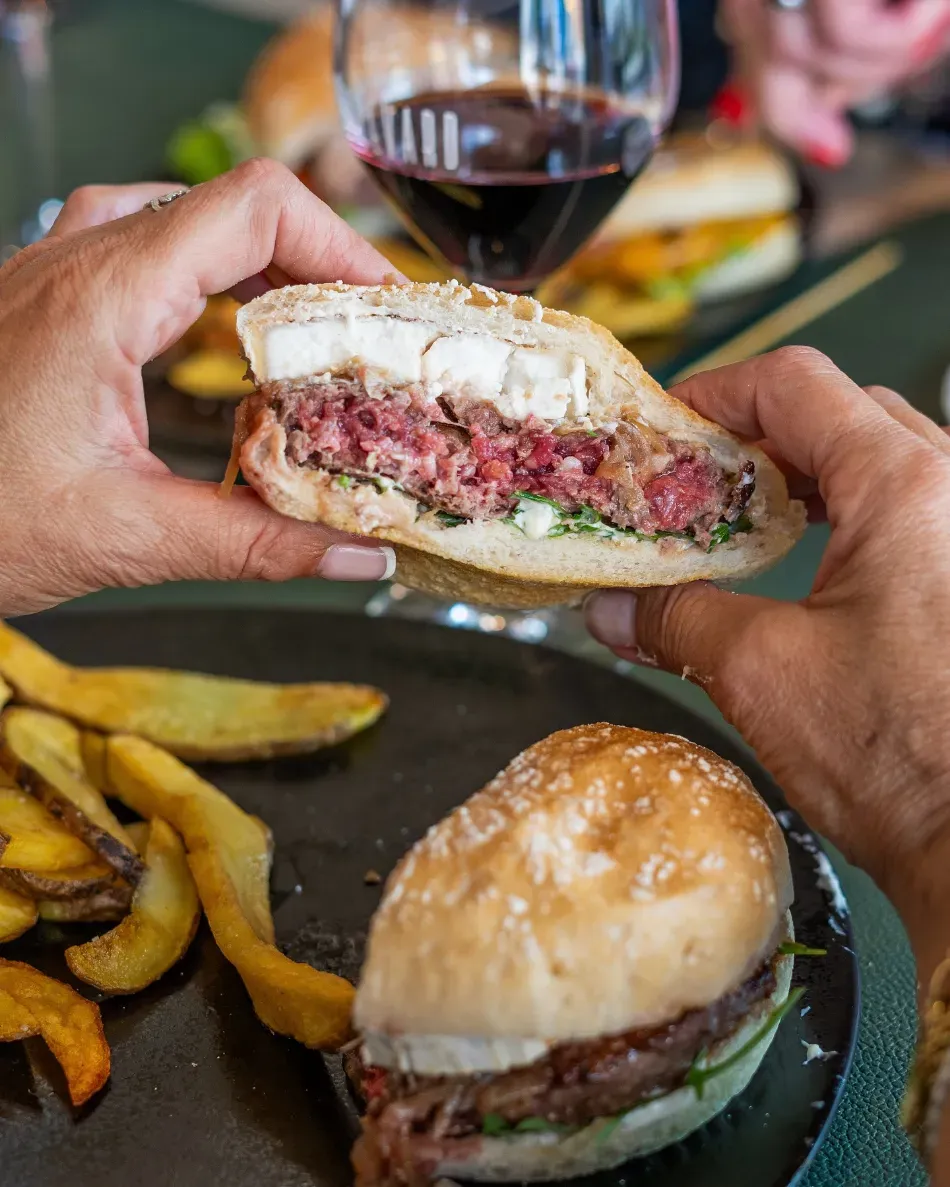 Unas manos sostienen una hamburguesa a medio comer con patatas fritas y una copa de vino tinto sobre una mesa.