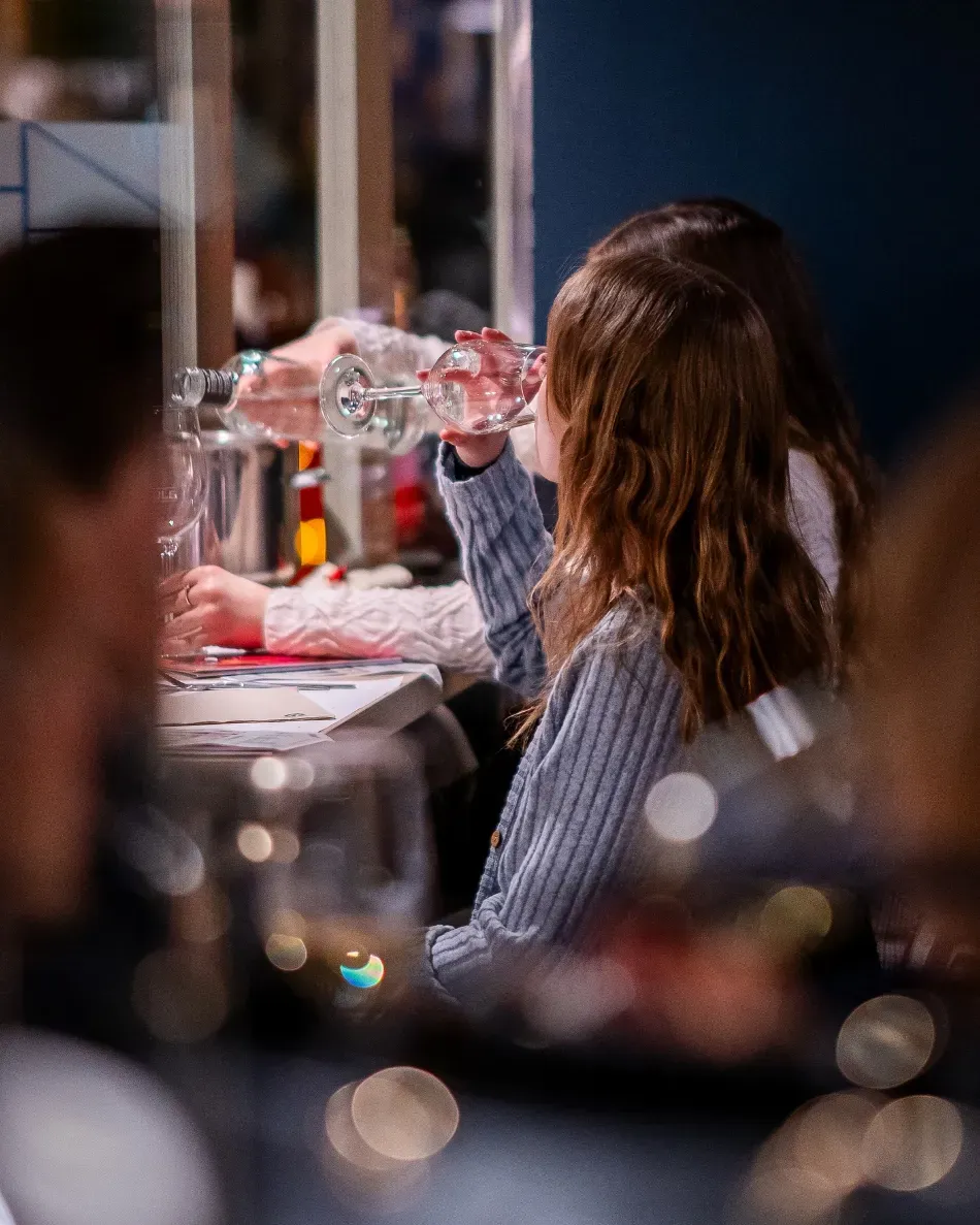 Una persona con una camisa a rayas bebe de una botella transparente en una mesa en un ambiente interior con poca luz.