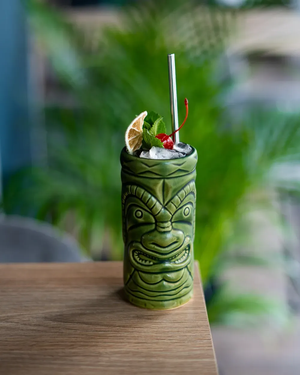 Cóctel en taza tiki verde con pajita y menta sobre una mesa de madera, plantas borrosas al fondo.