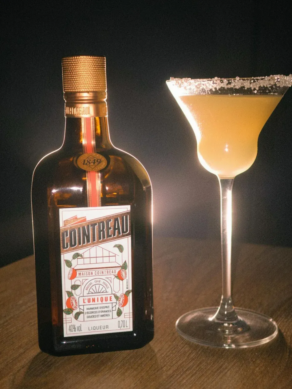 Botella de Cointreau junto a un cóctel de naranja espumoso en una copa de martini sobre un fondo oscuro.