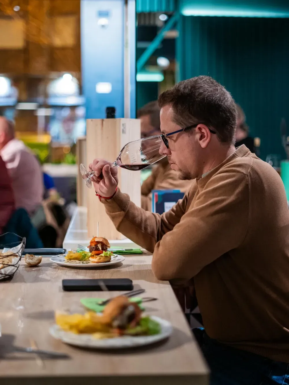 Un hombre con gafas degusta vino en la mesa de un restaurante con un plato servido y comensales borrosos al fondo.