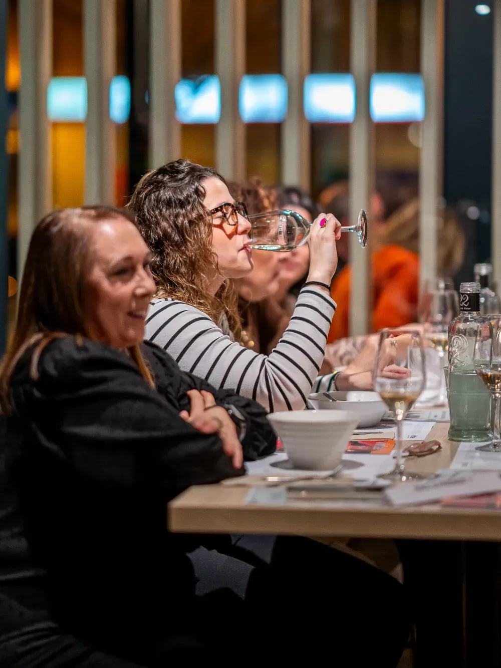Dos personas sentadas en la mesa de un restaurante; una bebe de un vaso mientras la otra observa.