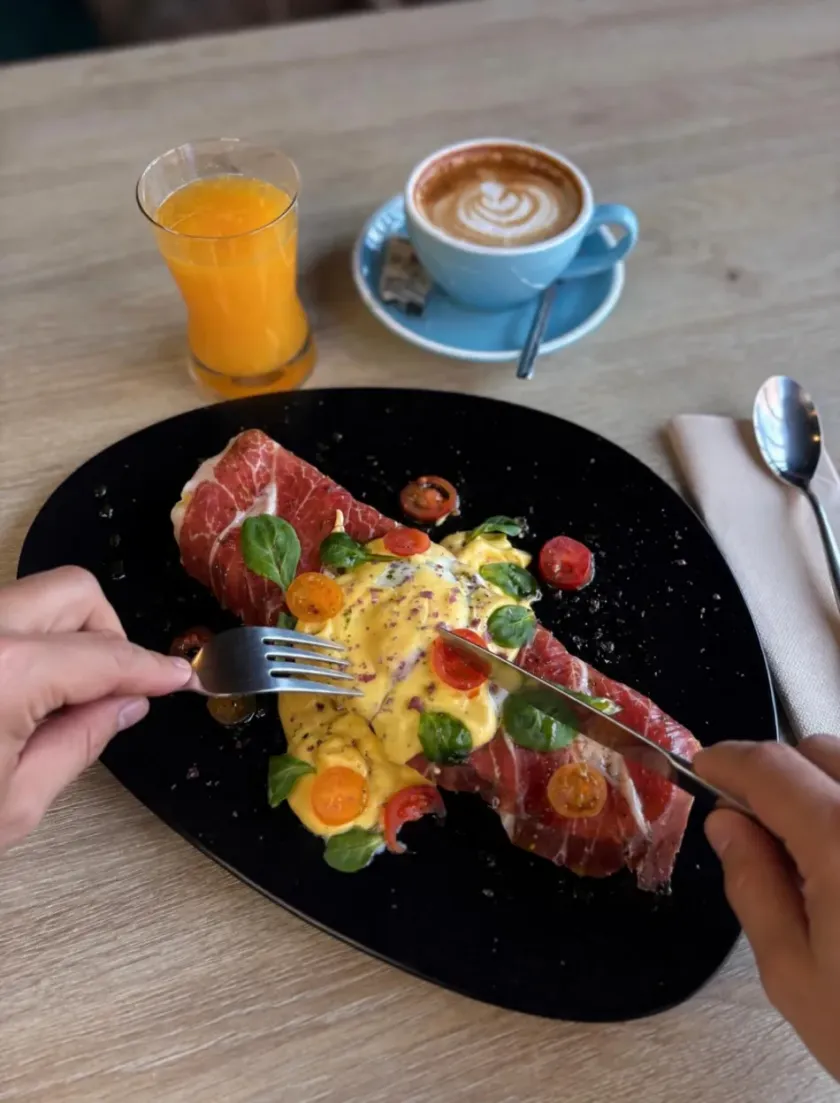 Manos comiendo un colorido plato de brunch con huevos, jamón, fruta, jugo de naranja y café sobre una mesa.