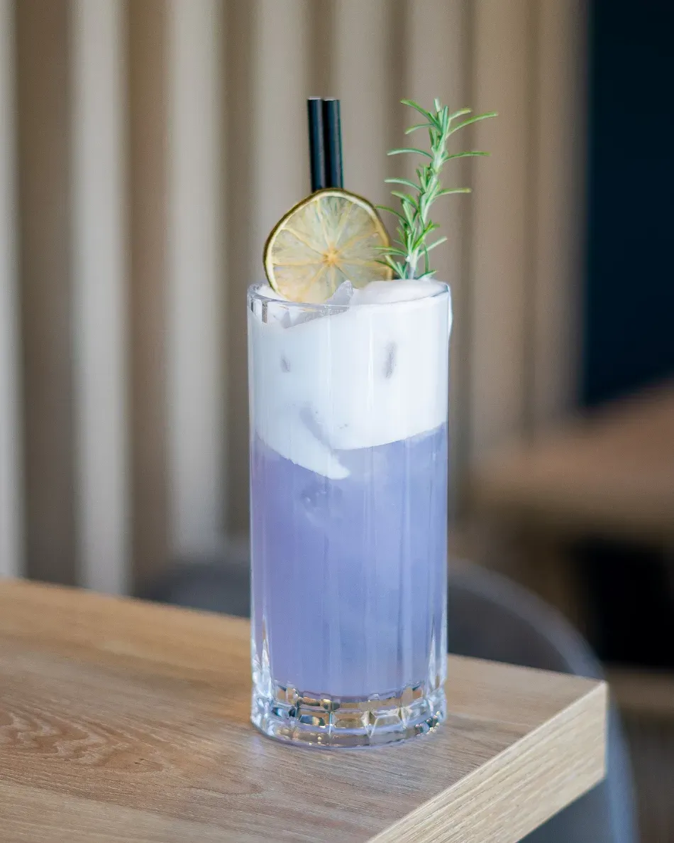 Cóctel de lavanda en un vaso alto con hielo, rodaja de lima, romero y una pajita negra sobre una mesa de madera.
