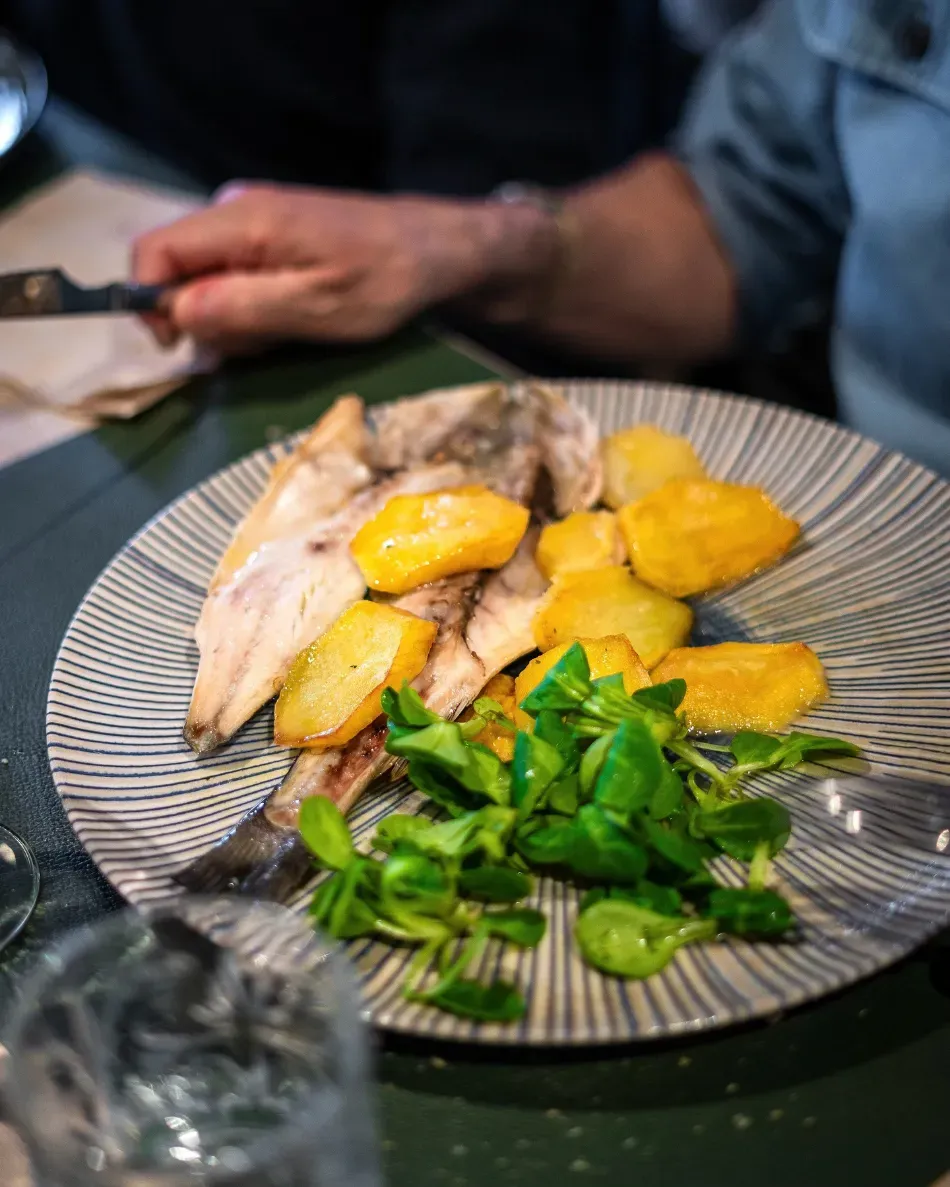 Pescado emplatado con trozos de mango amarillo y ensalada verde sobre una mesa, con la mano de una persona cerca.