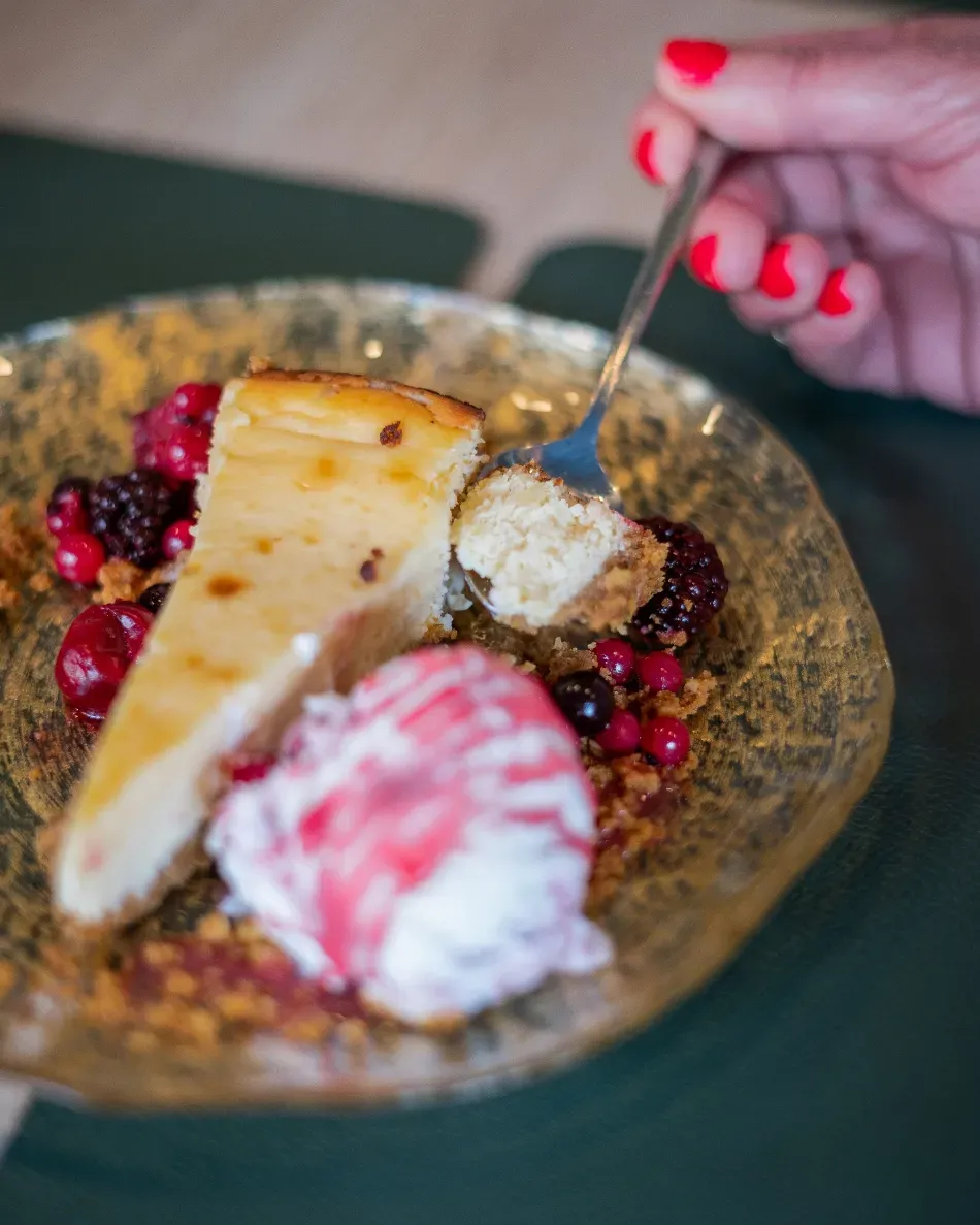Plato de postre con rebanadas de tarta de queso, bayas y una mano sosteniendo un tenedor sobre el plato.