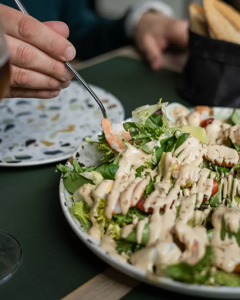 Una mano sosteniendo un tenedor sobre una ensalada con aderezo cremoso en un plato en una mesa de comedor.
