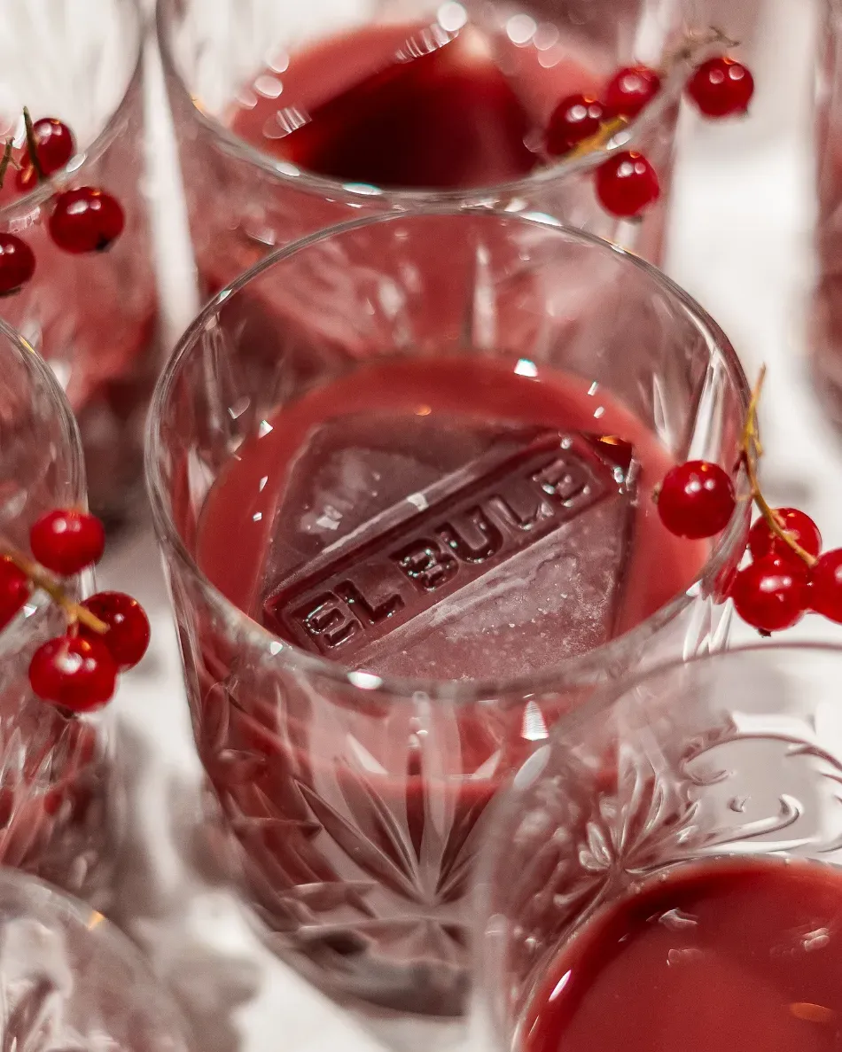 Vasos de cristal llenos de bebida de frutos rojos, adornados con grosellas rojas y un cubo de hielo con la etiqueta “BLEU”.