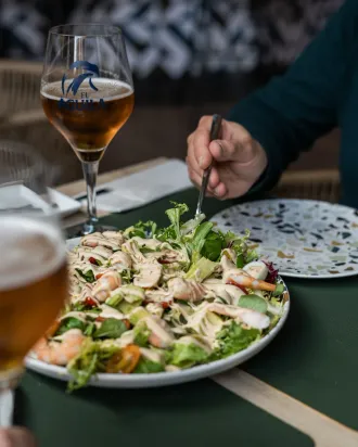 Ensalada sobre la mesa de un restaurante con una copa de vino y la mano de una persona que se introduce en ella con un tenedor.