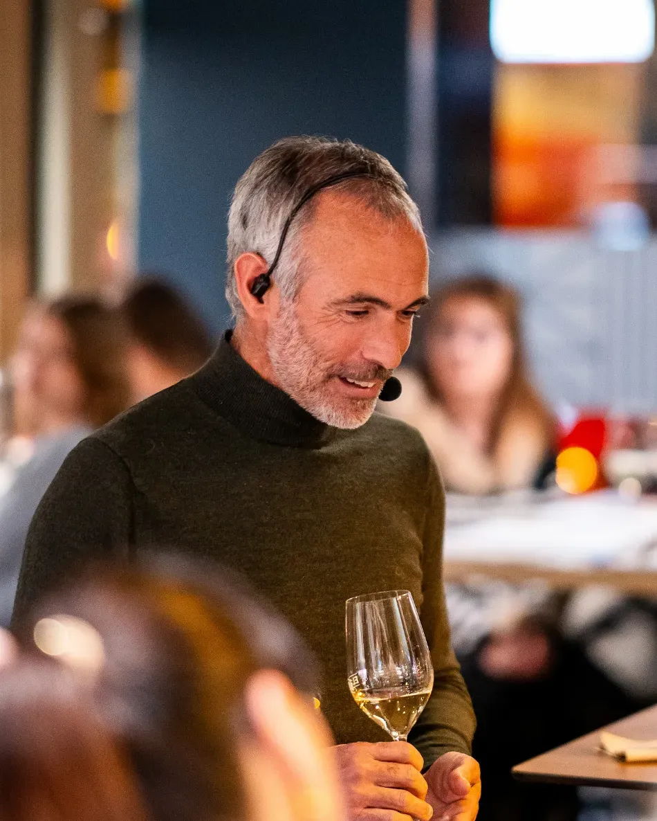 Hombre con suéter oscuro sosteniendo una copa de vino en la mesa de un restaurante, con comensales borrosos al fondo.