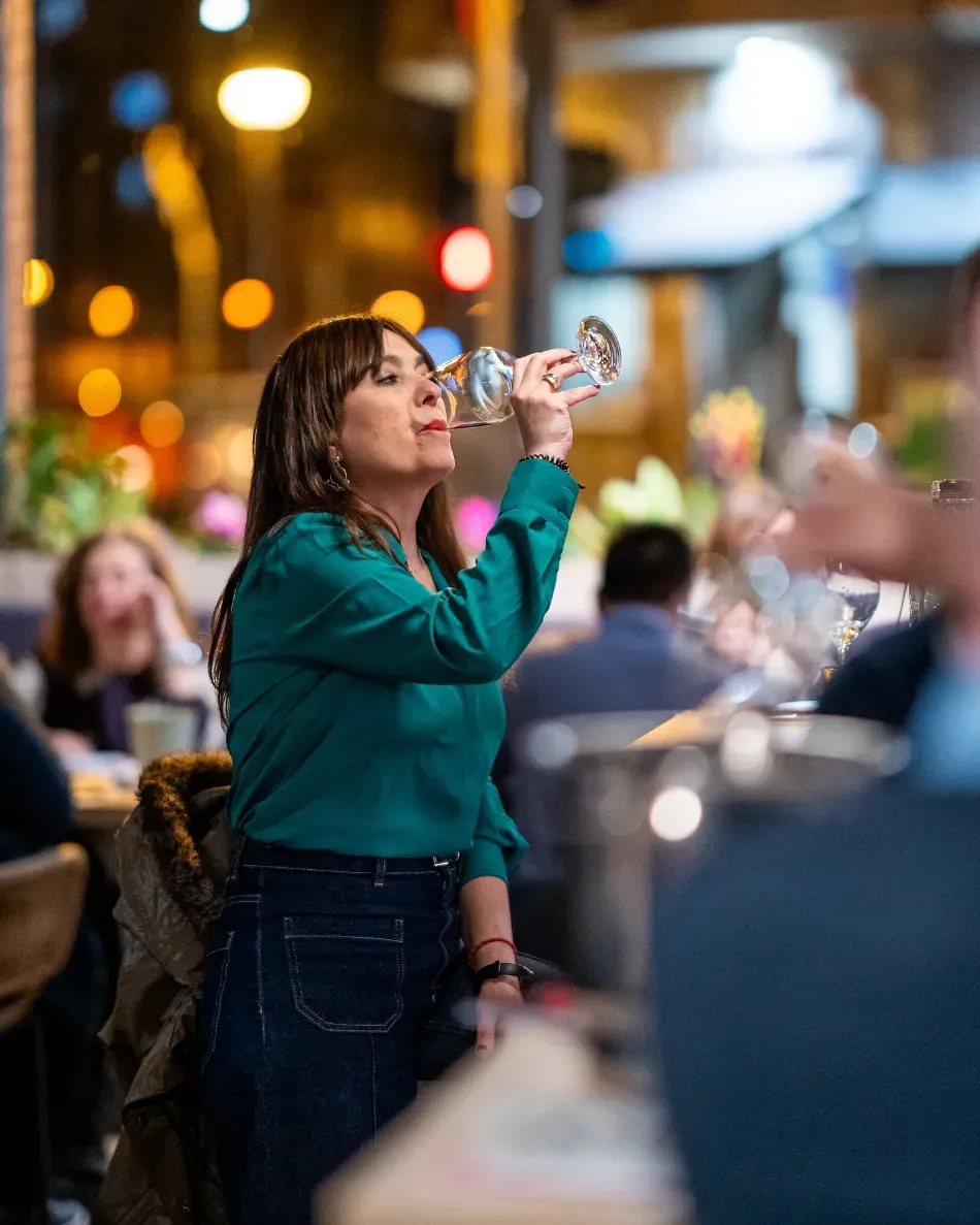 Una mujer con una chaqueta color turquesa bebe de una botella en una cafetería al aire libre con las luces borrosas de la ciudad de fondo.