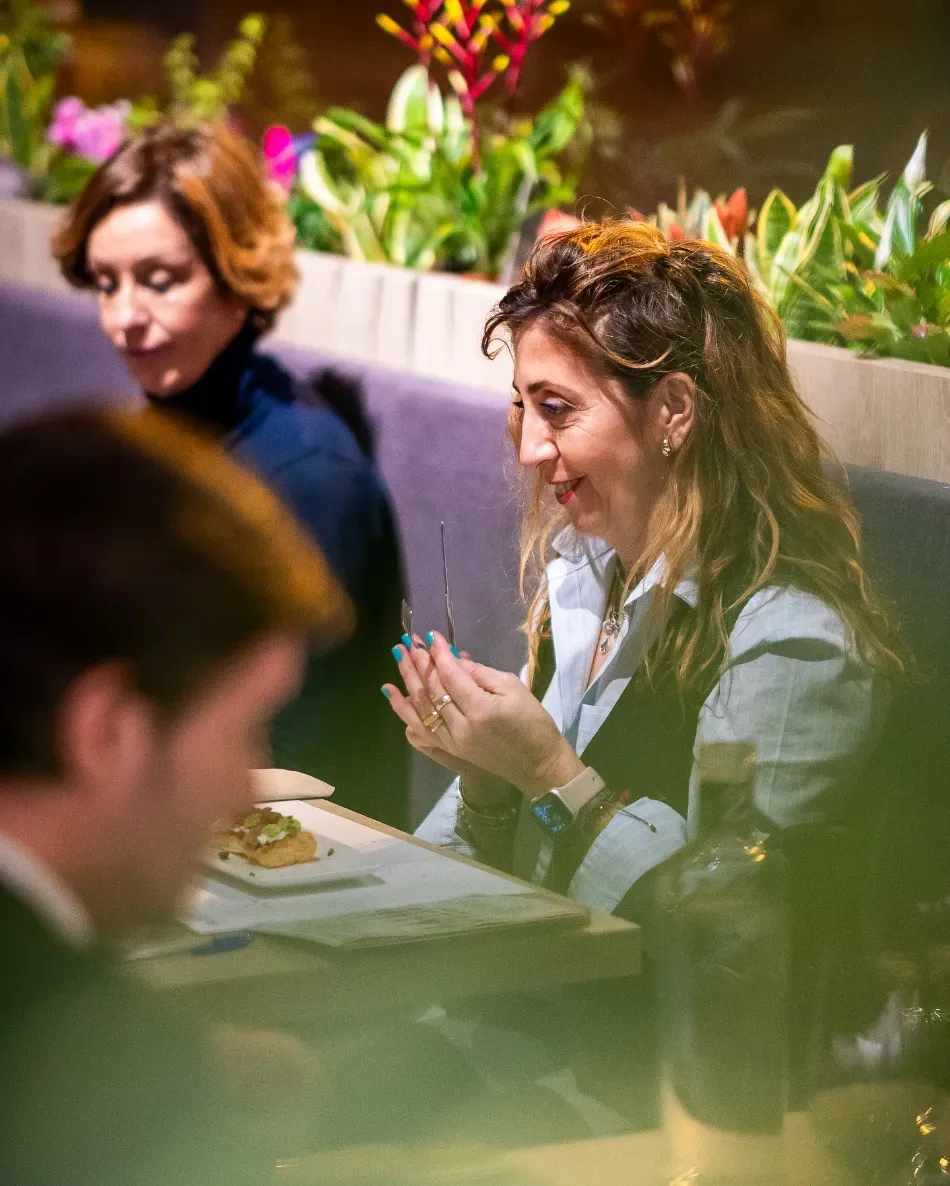 Varias personas estaban sentadas a una mesa en una sala de reuniones con iluminación tenue, donde una mujer hablaba y gesticulaba.