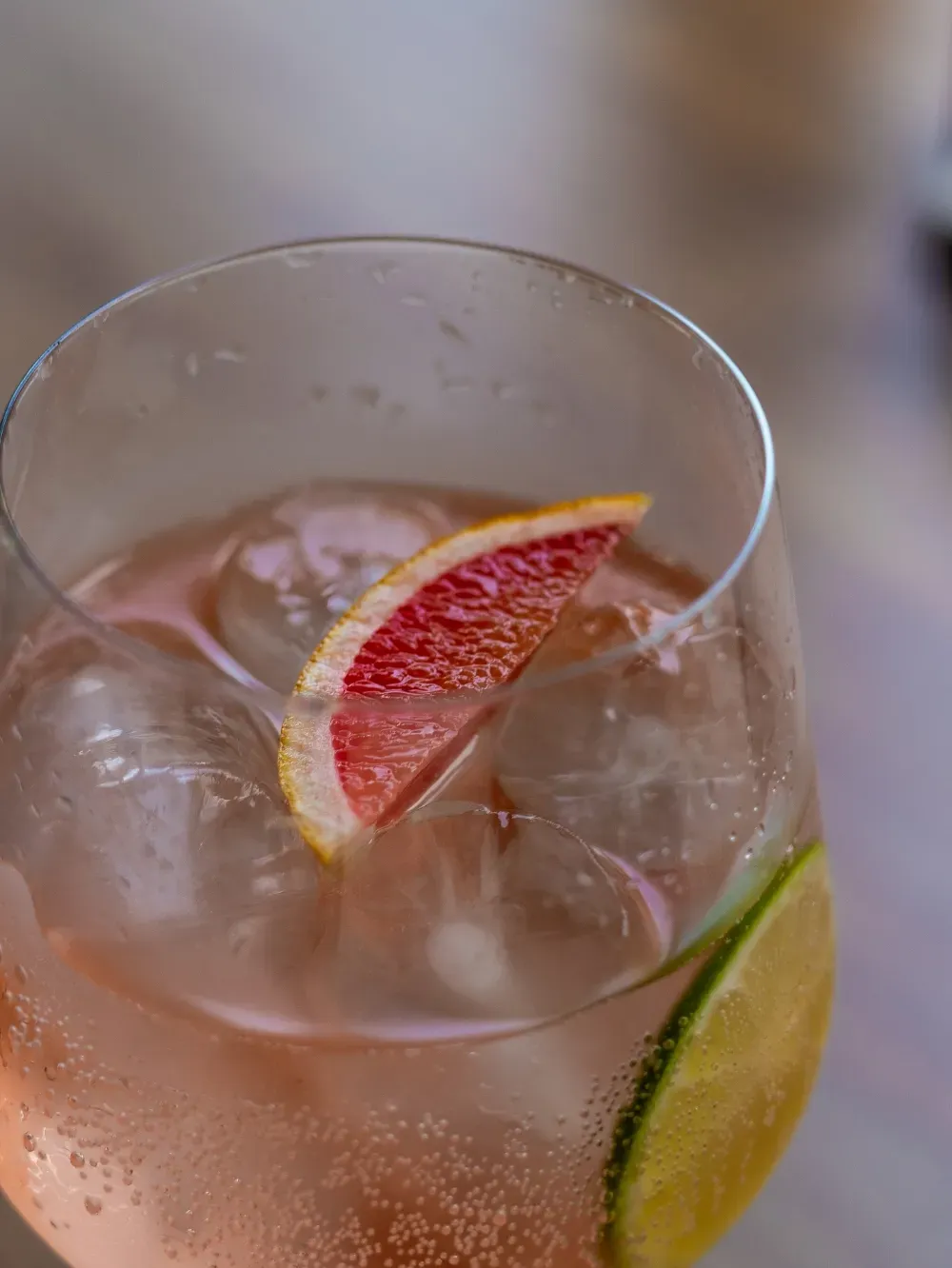 Primer plano de un cóctel rosa con hielo, una rodaja de pomelo y una rodaja de lima en un vaso transparente.