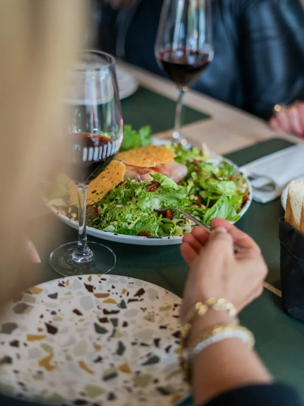 Mesa de restaurante con ensalada, pan, copas de vino y una persona extendiendo la mano para coger comida.