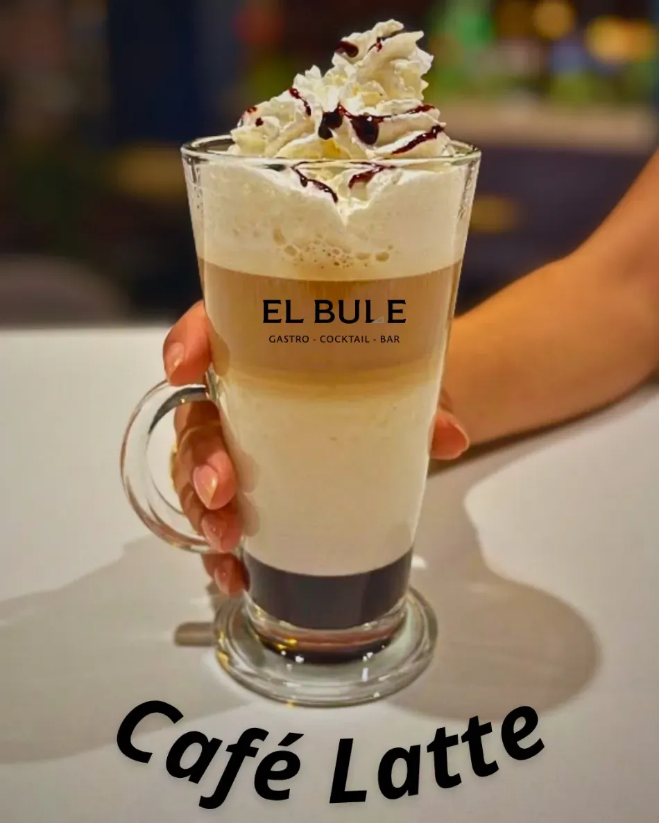 Café con leche cremoso en una taza de cristal con nata montada, sostenido sobre una mesa con la etiqueta 