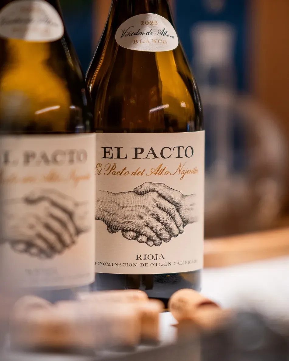 Dos botellas de vino El Pacto con etiquetas beige sobre una mesa, con los corchos en primer plano.