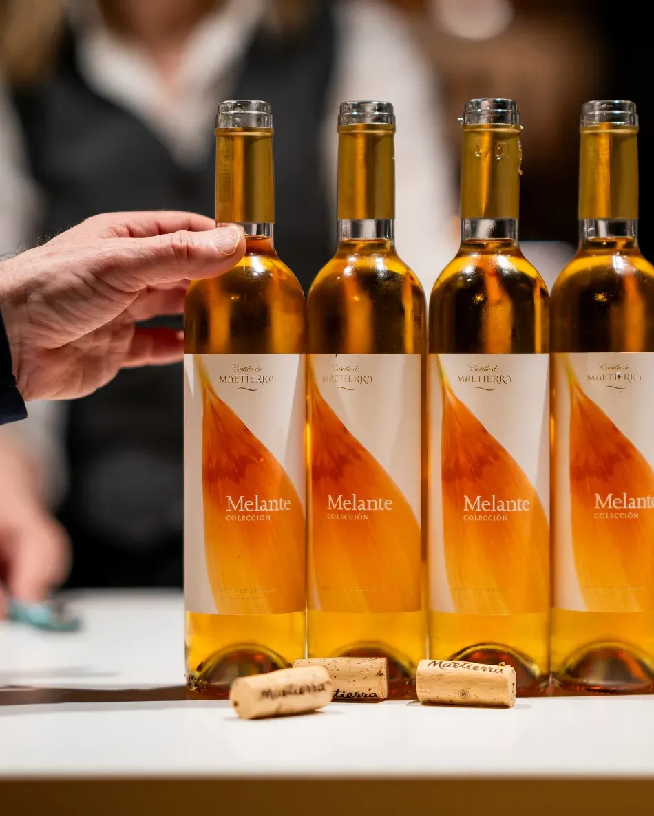 Cuatro botellas de vino ámbar con etiquetas blancas sobre una mesa, con una mano extendiéndose hacia ellas.