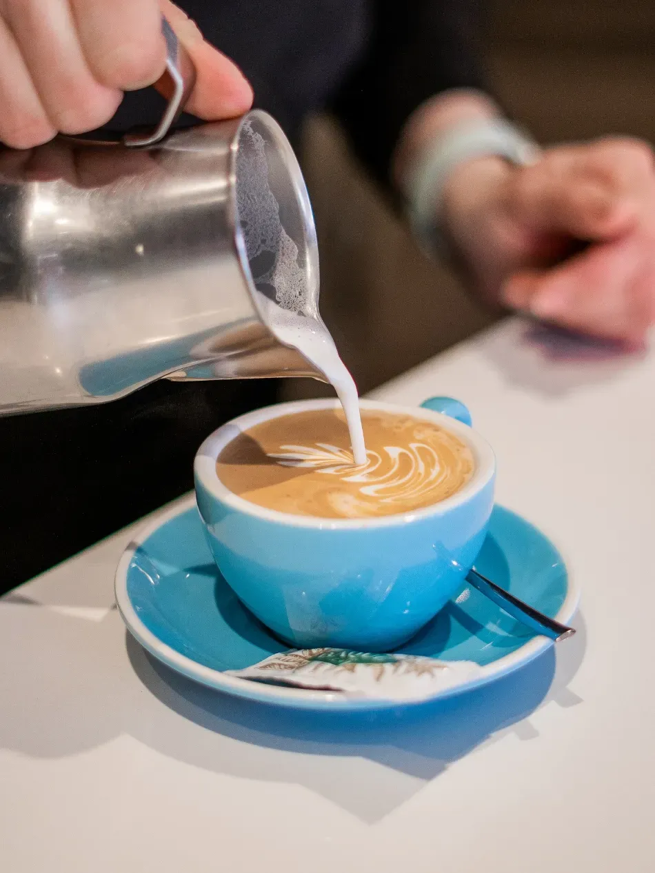 Vertiendo arte latte en una taza azul sobre un platillo en una mesa de café.
