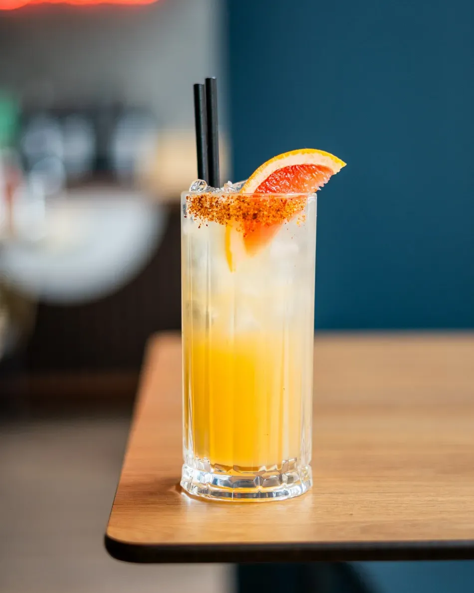 Cóctel alto de naranja con guarnición de pomelo sobre una mesa de madera, con pajitas negras y un fondo desenfocado.