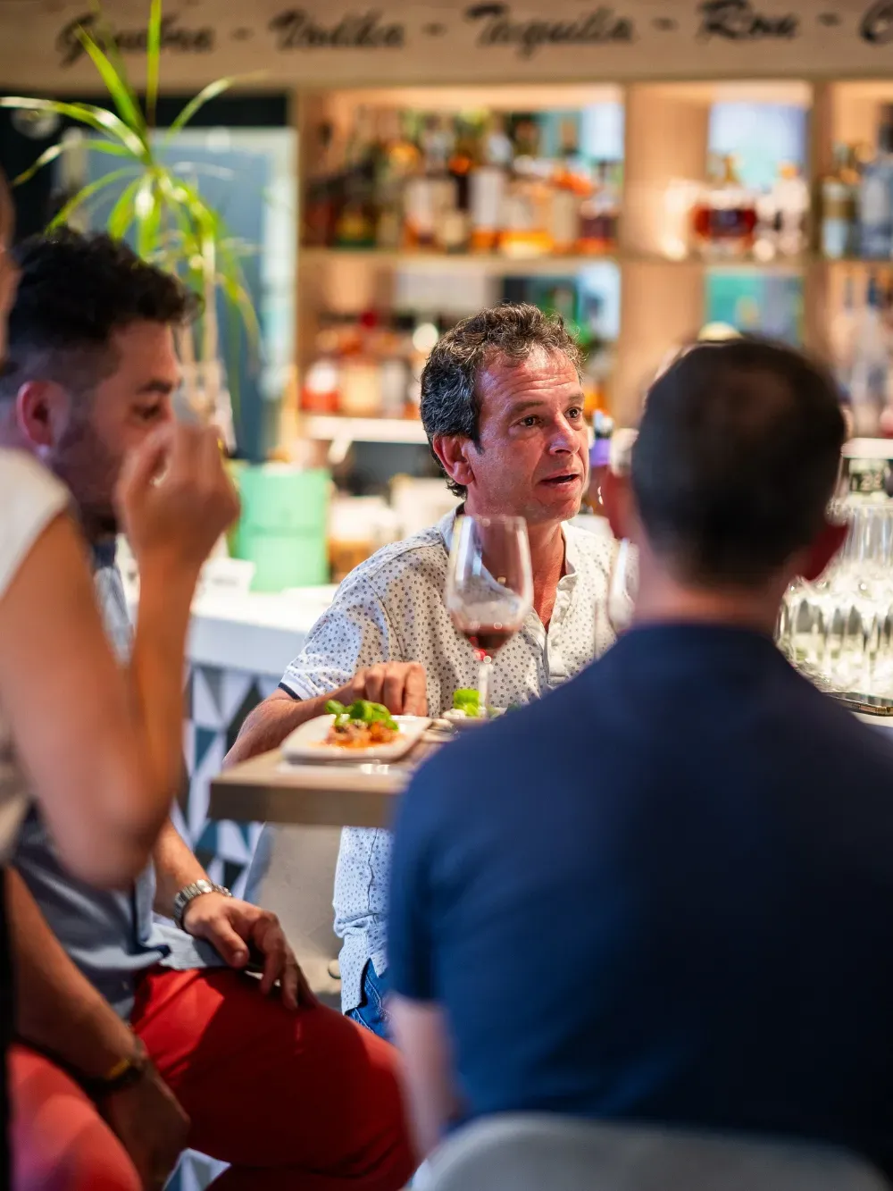 Personas sentadas en una mesa de café, un hombre hablando mientras otros escuchan, con bebidas y comida en un ambiente cálido y acogedor.