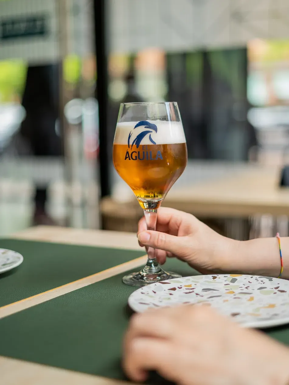Una mano sostiene una copa de cerveza con tallo sobre una mesa en un ambiente informal de cafetería al aire libre.