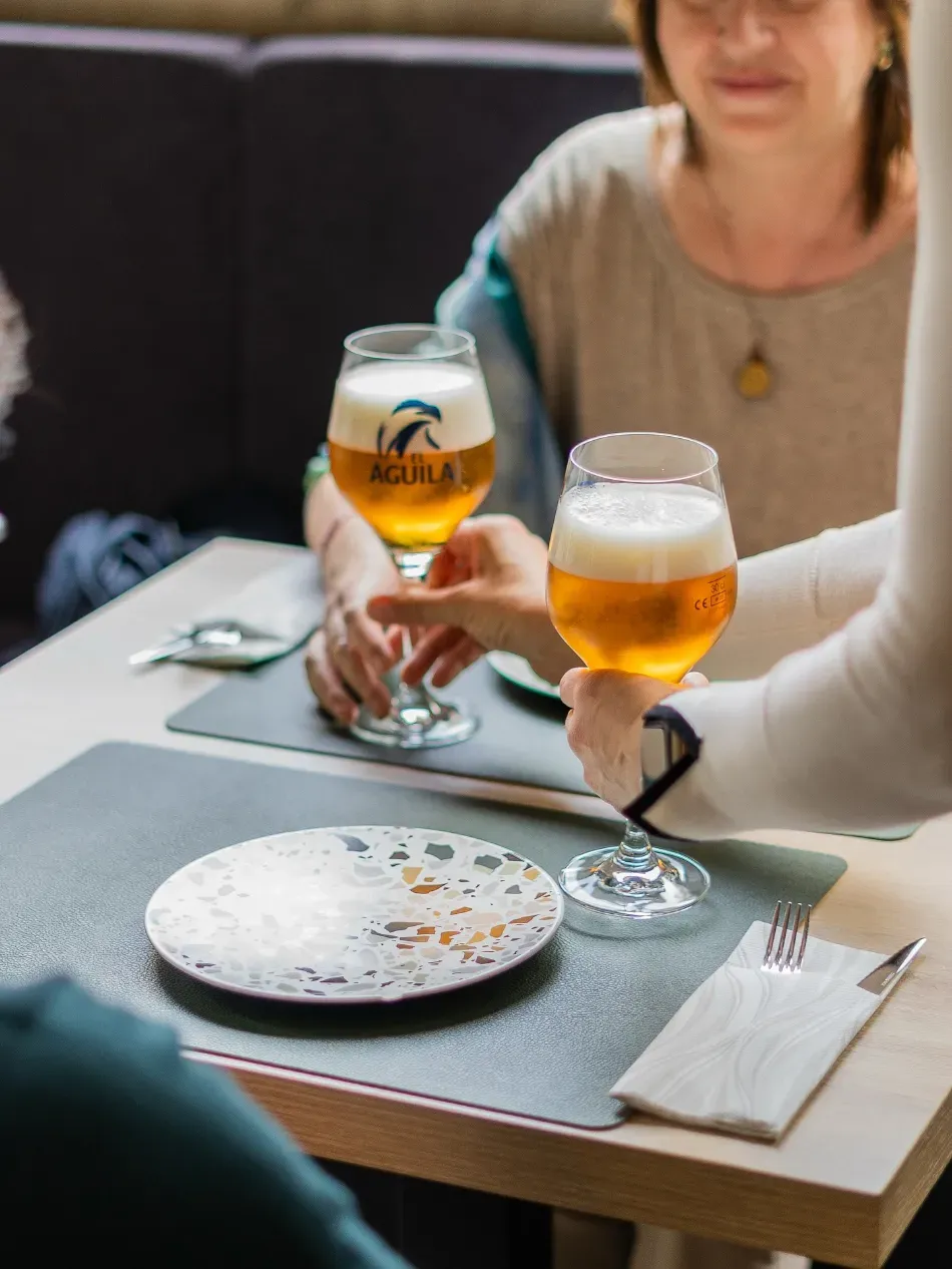 Dos personas brindan con vasos de cerveza en una mesa, con un plato vacío y servilletas en un restaurante acogedor.