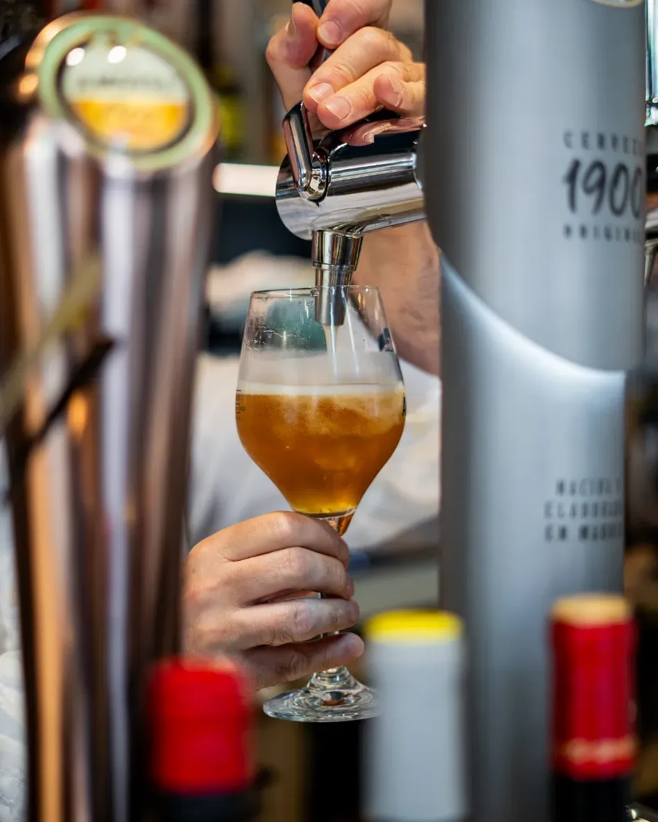 Vertiendo cerveza a mano desde un grifo en un vaso, con botellas borrosas en primer plano y un bar de fondo.