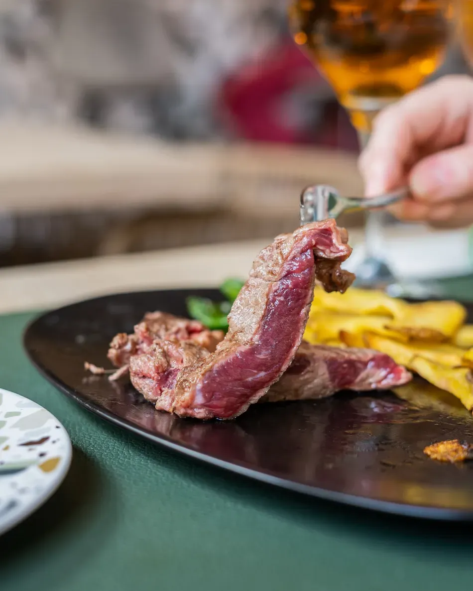 Filete en un plato negro con patatas asadas, sostenido con un tenedor en la mesa de un restaurante.