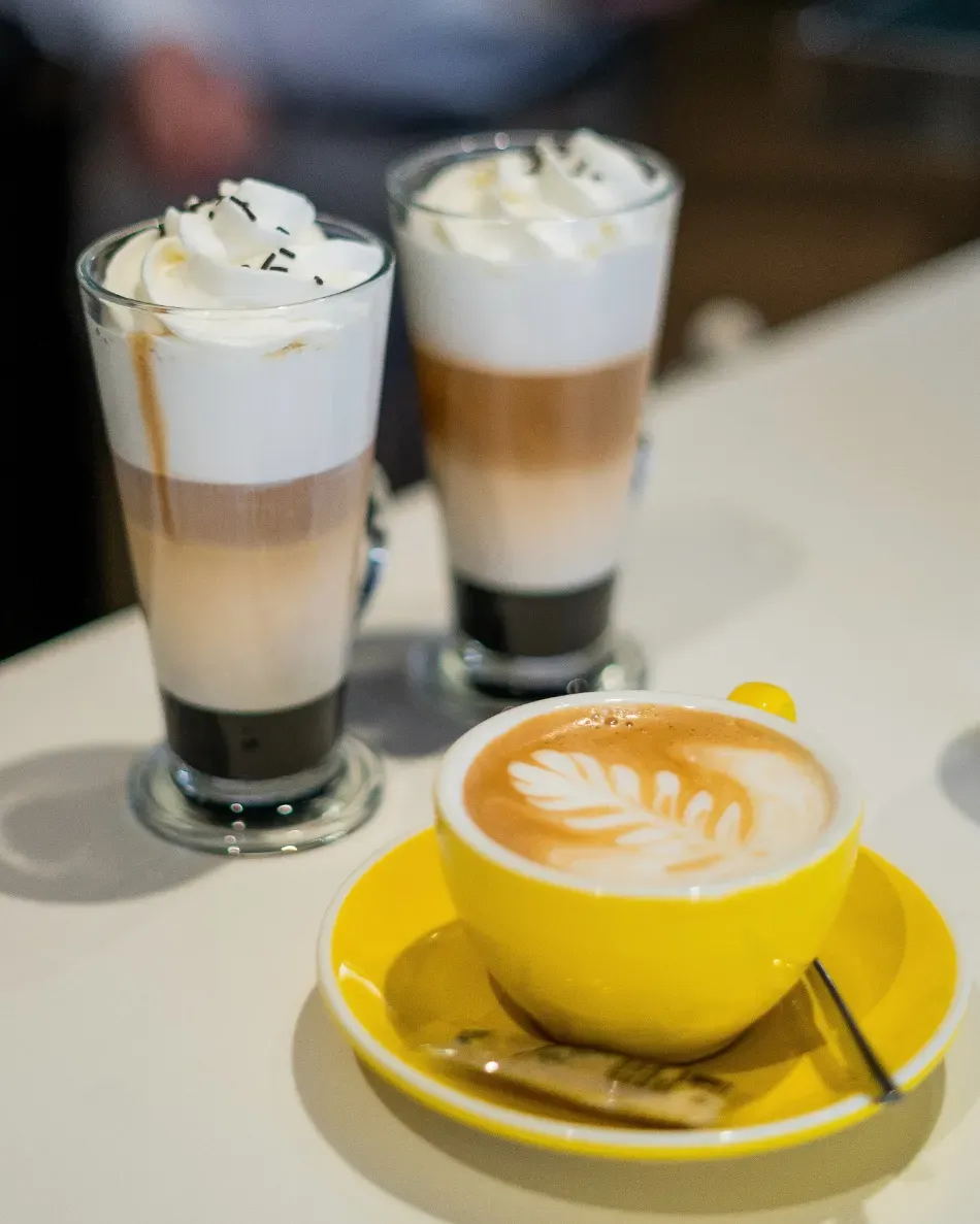 Dos bebidas de café en capas con crema batida junto a una taza de capuchino amarilla sobre una mesa.
