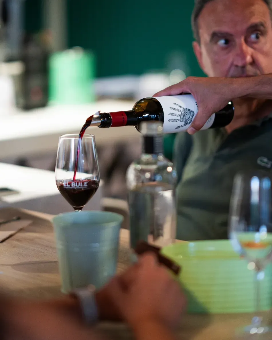 Se sirve vino en una copa sobre una mesa, con botellas y copas de colores en primer plano.