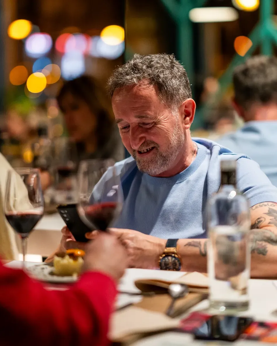 Hombre con camisa azul claro usando un teléfono inteligente en la mesa de un restaurante con bebidas y un fondo bokeh iluminado con velas.