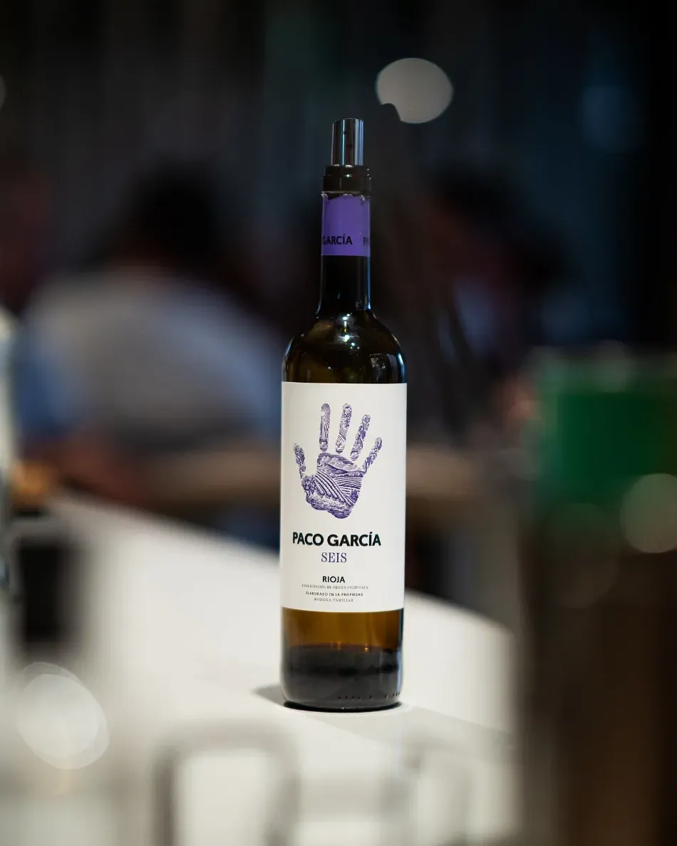 Botella de vino con etiqueta morada y huella de mano sobre un pequeño pedestal blanco, sobre un fondo oscuro difuminado.
