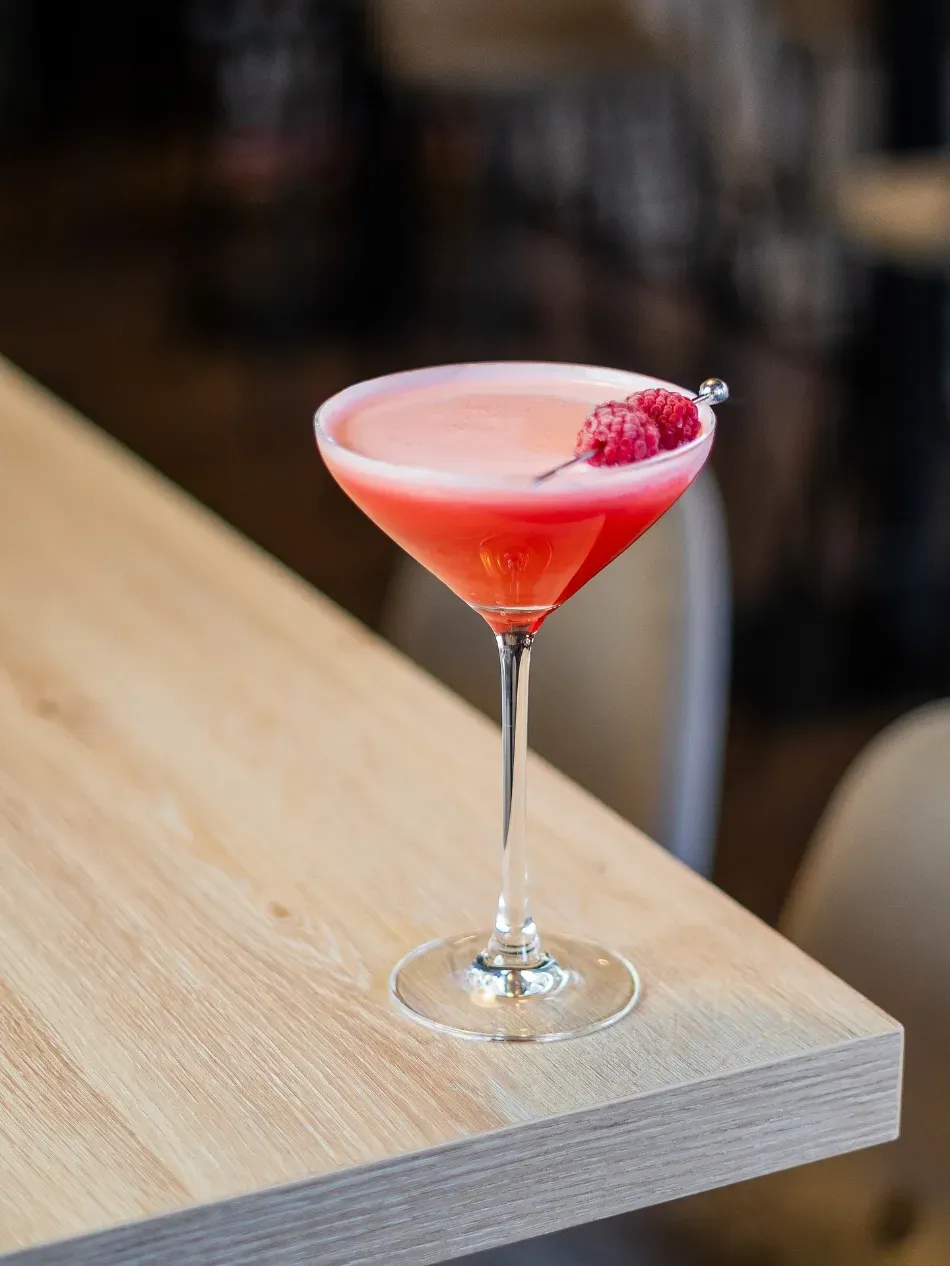 Cóctel rosa en una copa de martini con una frambuesa sobre una mesa de madera.