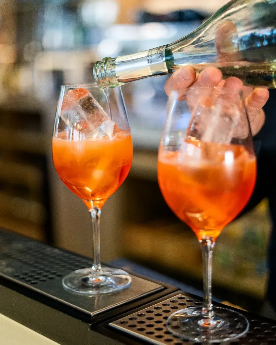Dos cócteles de naranja siendo servidos de una botella en copas con tallo en un bar.