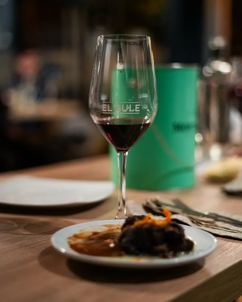 Una copa de vino junto a un postre emplatado en la mesa de un restaurante, con un fondo de comedor desenfocado.
