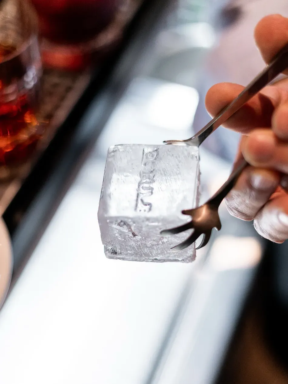 Una mano usa pinzas para sujetar un cubo de hielo transparente sobre la barra.