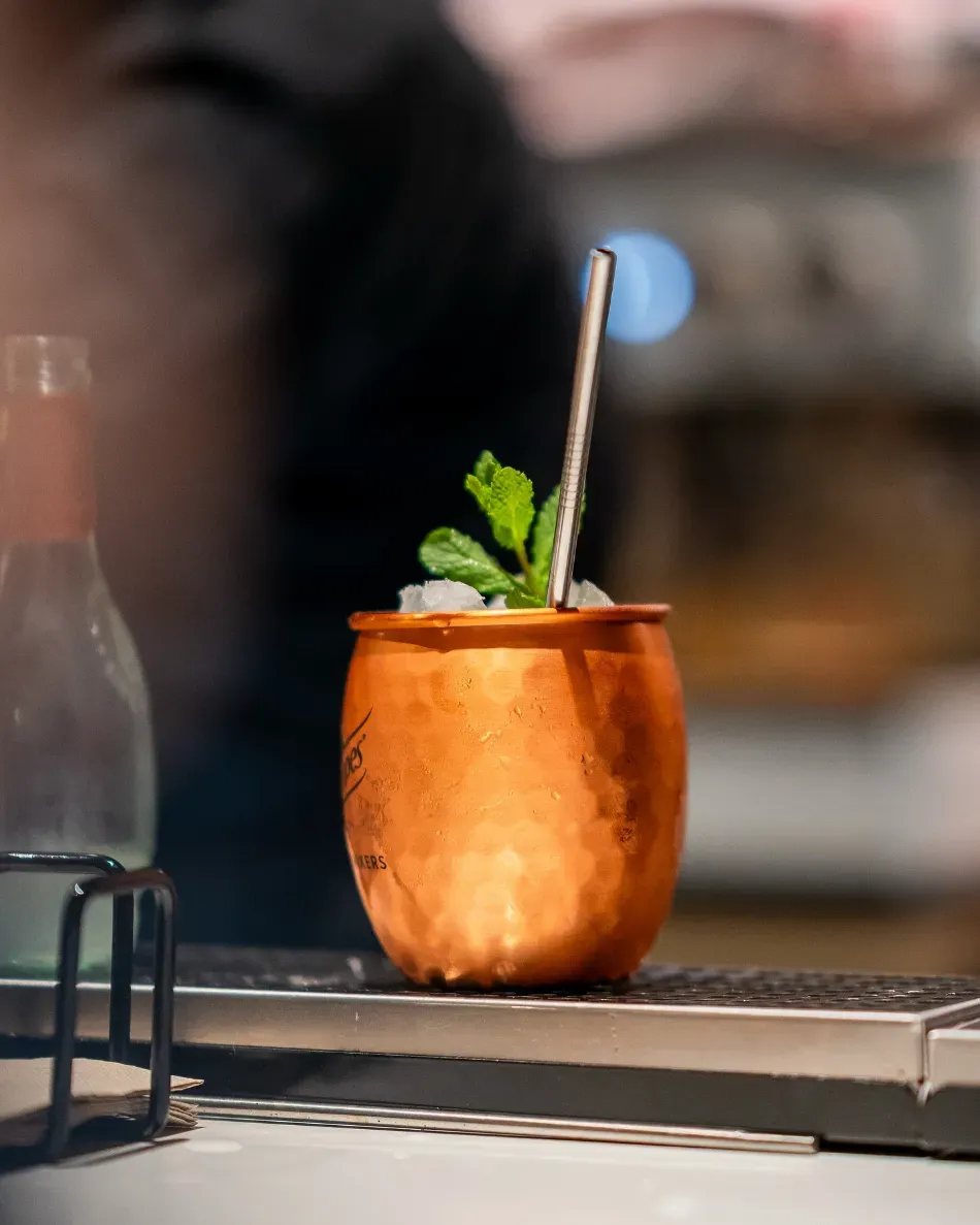 Cóctel en taza de cobre con menta y pajita sobre la barra de un bar, fondo desenfocado.