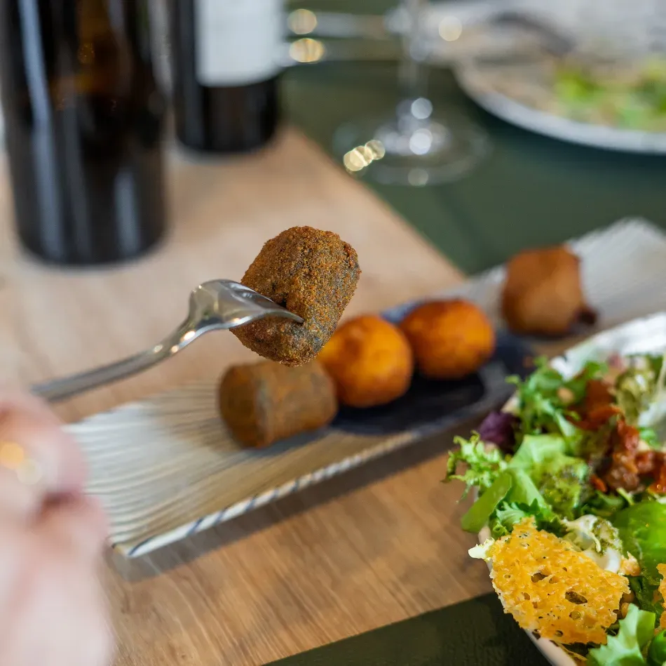 Tenedor con un aperitivo frito sobre una bandeja de bocadillos variados junto a una ensalada en una mesa de madera.