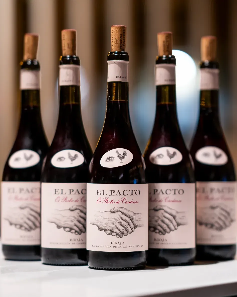 Fila de botellas de vino El Pacto con etiquetas blancas y corchos en un estante.