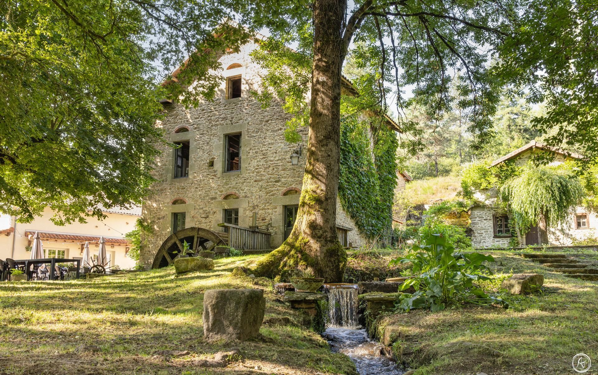 Ancien moulin en pierre avec roue à aubes, ruisseau et arbres environnants dans un cadre verdoyant et luxuriant.