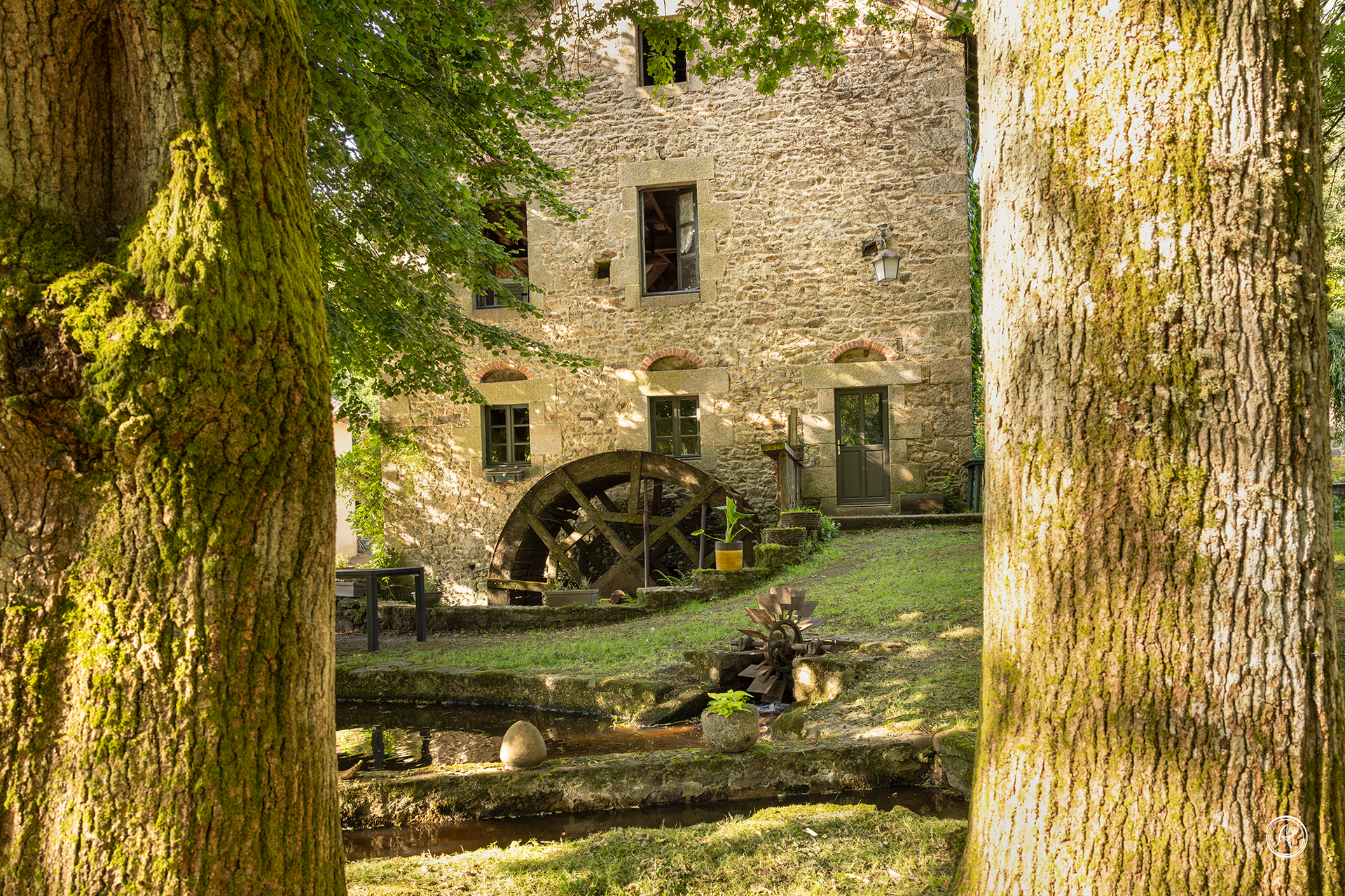 Bâtiment en pierre abritant un moulin à eau avec une roue à aubes en bois et des arbres.