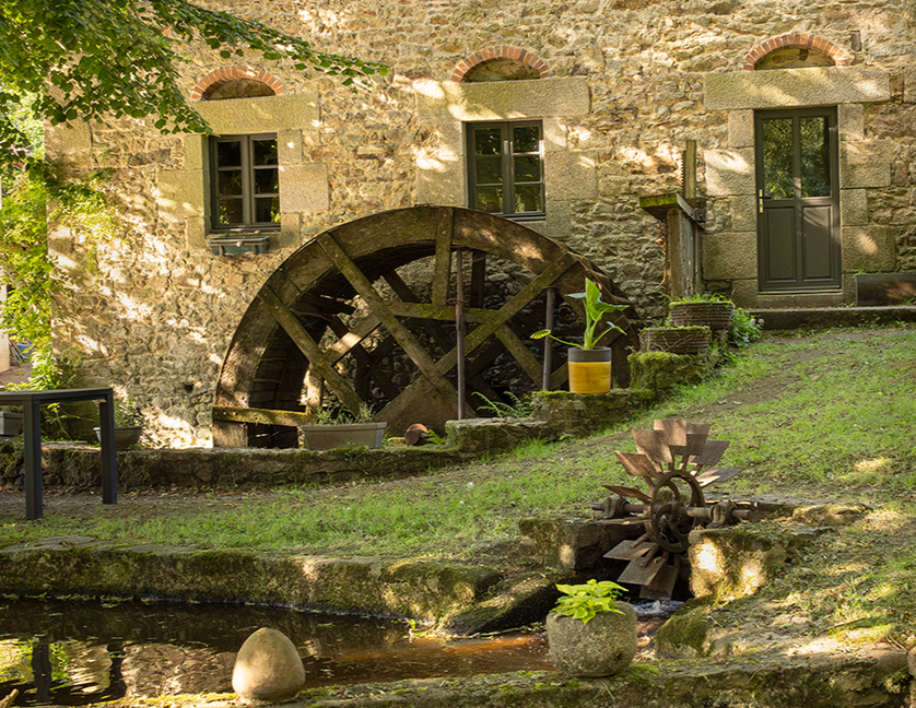 Une roue à aubes usée par le temps se dresse devant un bâtiment en pierre avec des fenêtres et une porte ; journée ensoleillée.