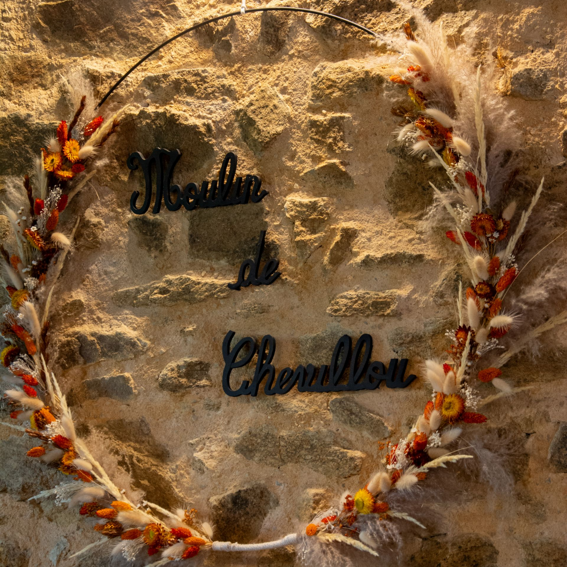 Une couronne de fleurs portant l'inscription Moulin de Chevillou est accrochée contre un mur de pierre.