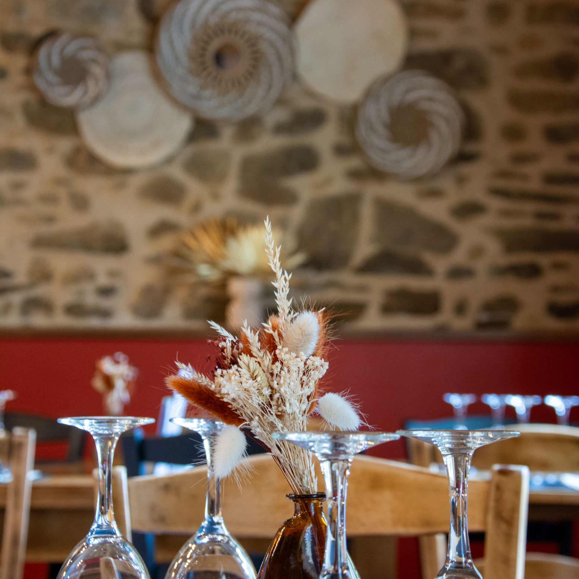 Dressage de table dans un restaurant avec verres à vin, fleurs séchées et décoration murale tissée, le tout adossé à un mur de pierre.