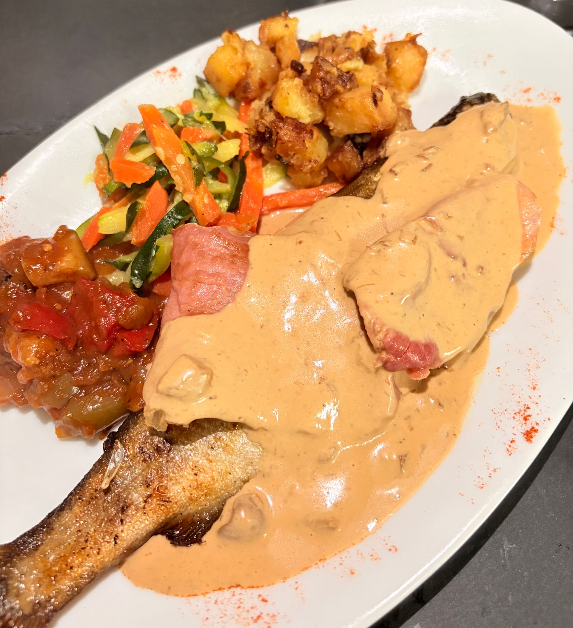 Un filet de poisson nappé d'une sauce crémeuse, servi avec des pommes de terre rôties, des légumes coupés en tranches et une portion de salsa rouge.