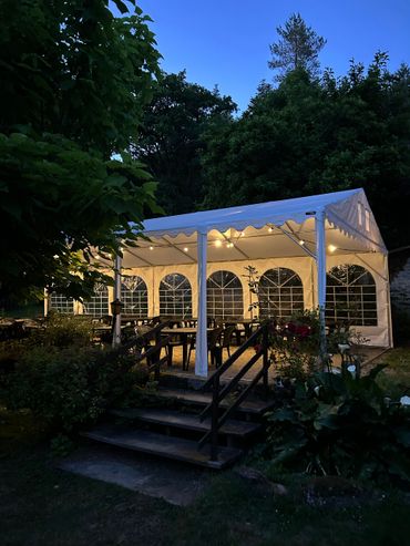 Tente extérieure à toit blanc avec tables et chaises, éclairée par des guirlandes lumineuses, située à côté d'un escalier et entourée d'arbres.
