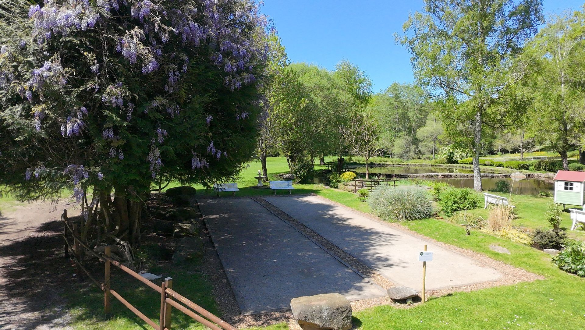 Camping avec places de parking en gravier, arbre en fleurs et petit étang dans un environnement verdoyant et ensoleillé.