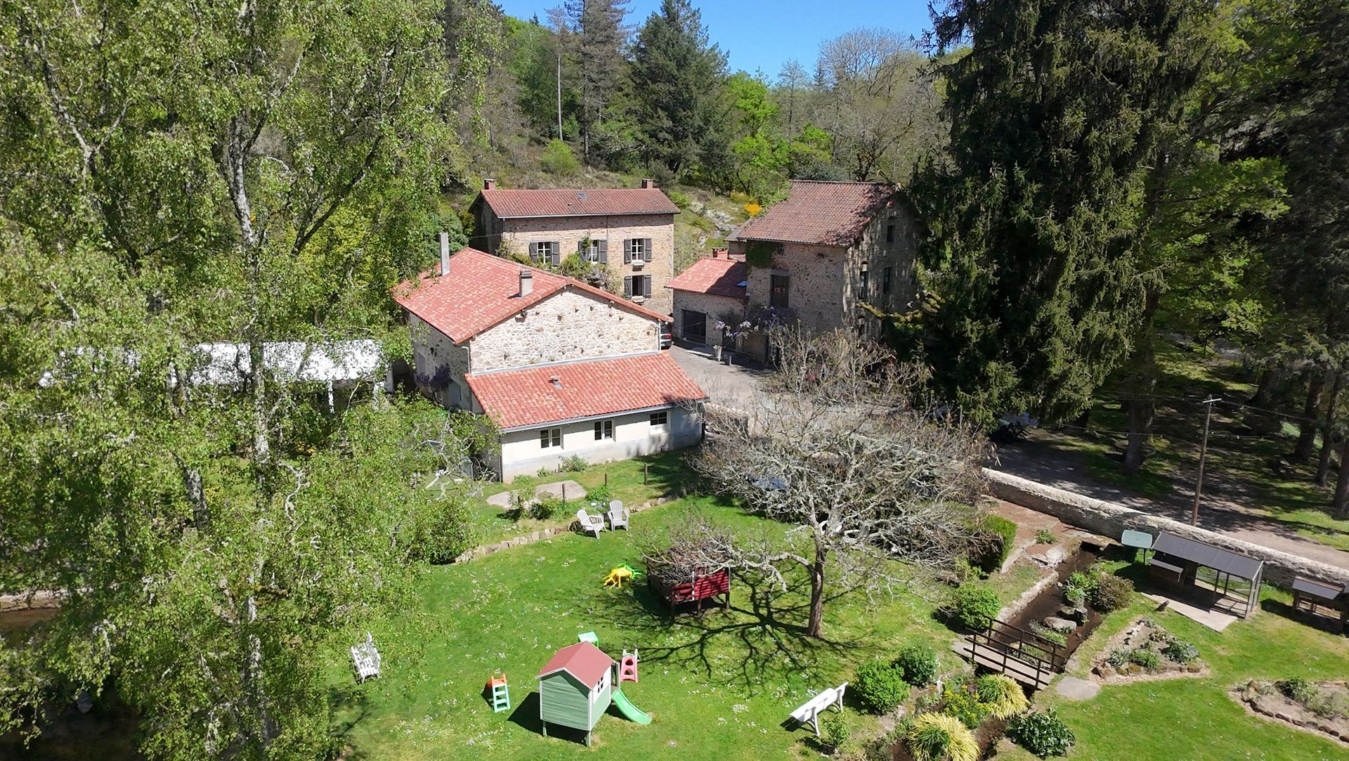 Des bâtiments en pierre aux toits de tuiles rouges nichés parmi les arbres, une pelouse verdoyante avec une maisonnette et un jardin.