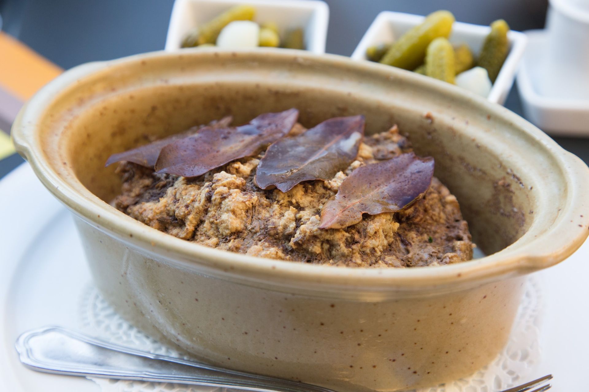 Plat de pâté garni de feuilles, servi avec des cornichons dans des petits bols.